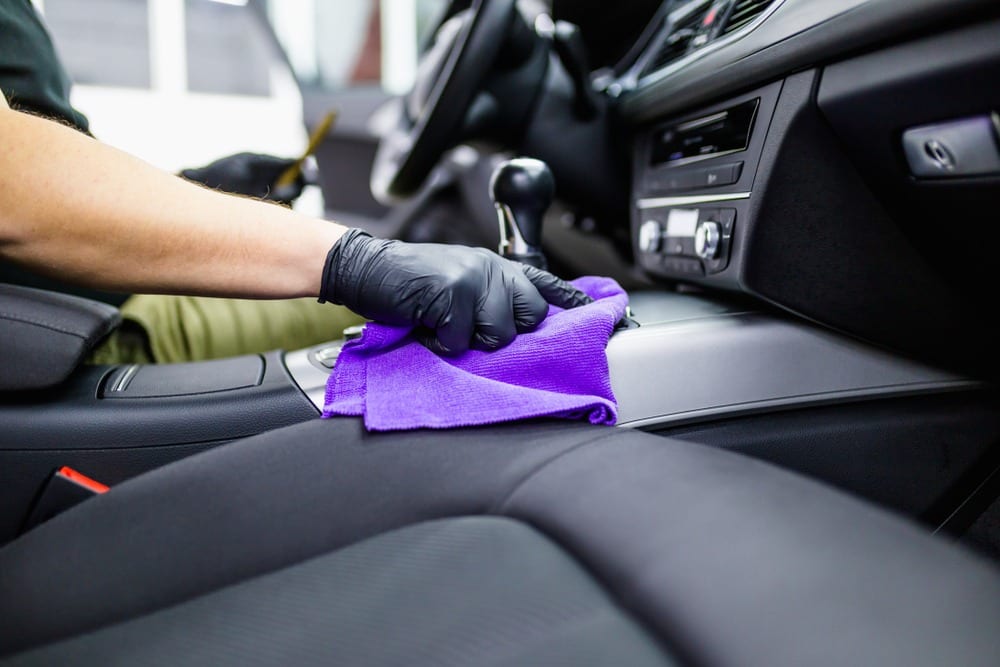 Person detailing a car's centre console with a purple microfibre cloth, focusing on detailed interior cleaning.