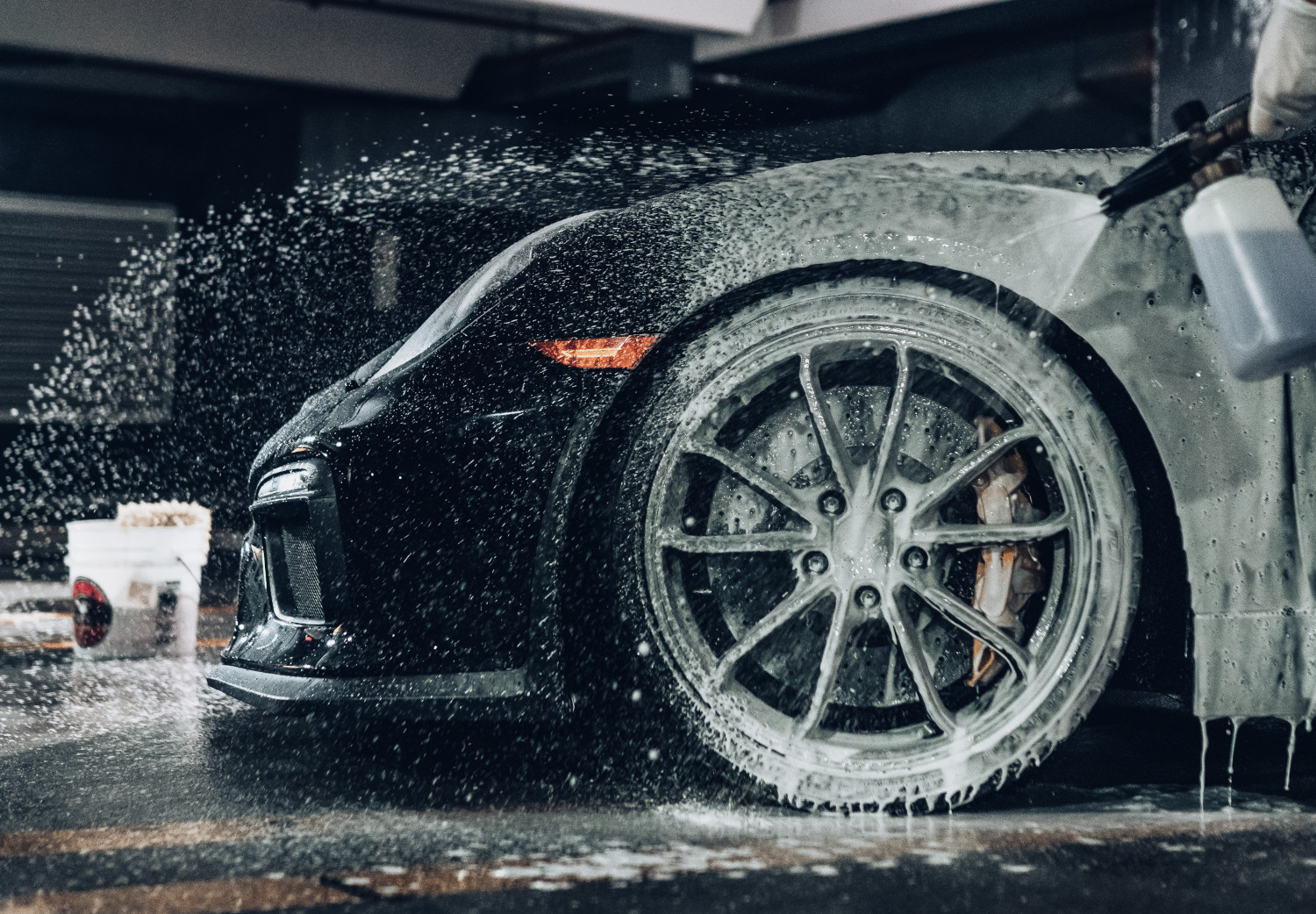 High-pressure spray cleaning the wheel and side of a black sports car in a self-service car wash setting.