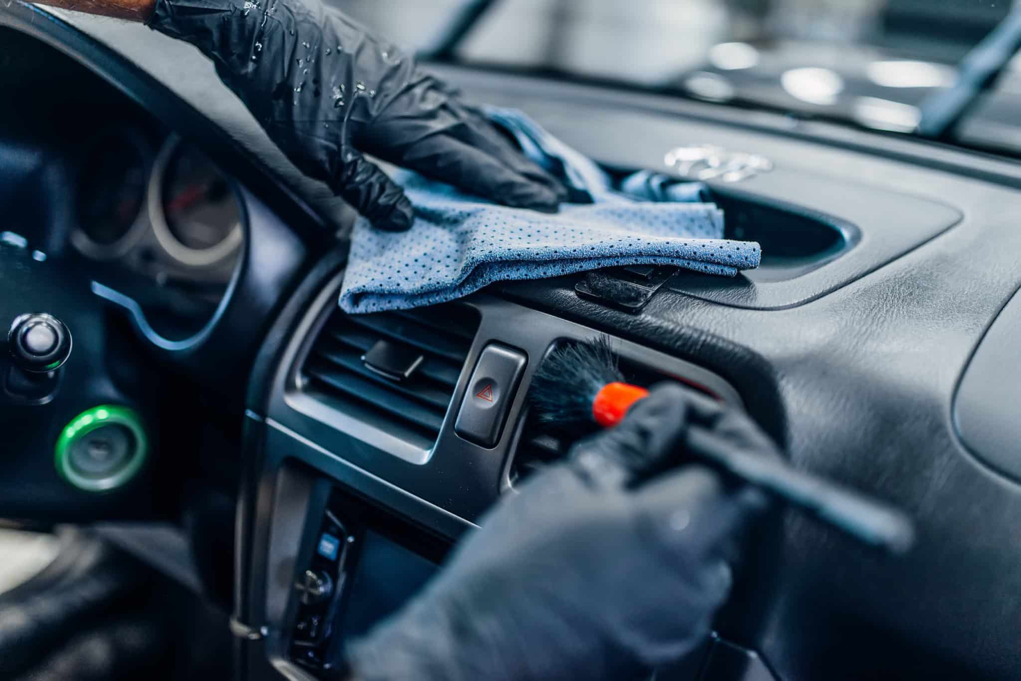 Close-up of a professionally cleaning a car's dashboard with a cloth and brush, focusing on the air vents and control buttons.