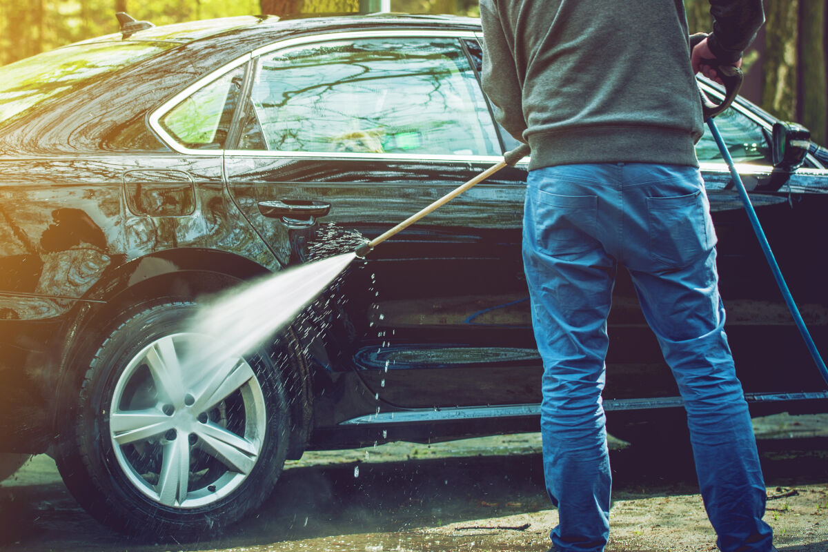 Person using a high-pressure hose and brush to wash a black sedan, focusing on the car's side and wheel.