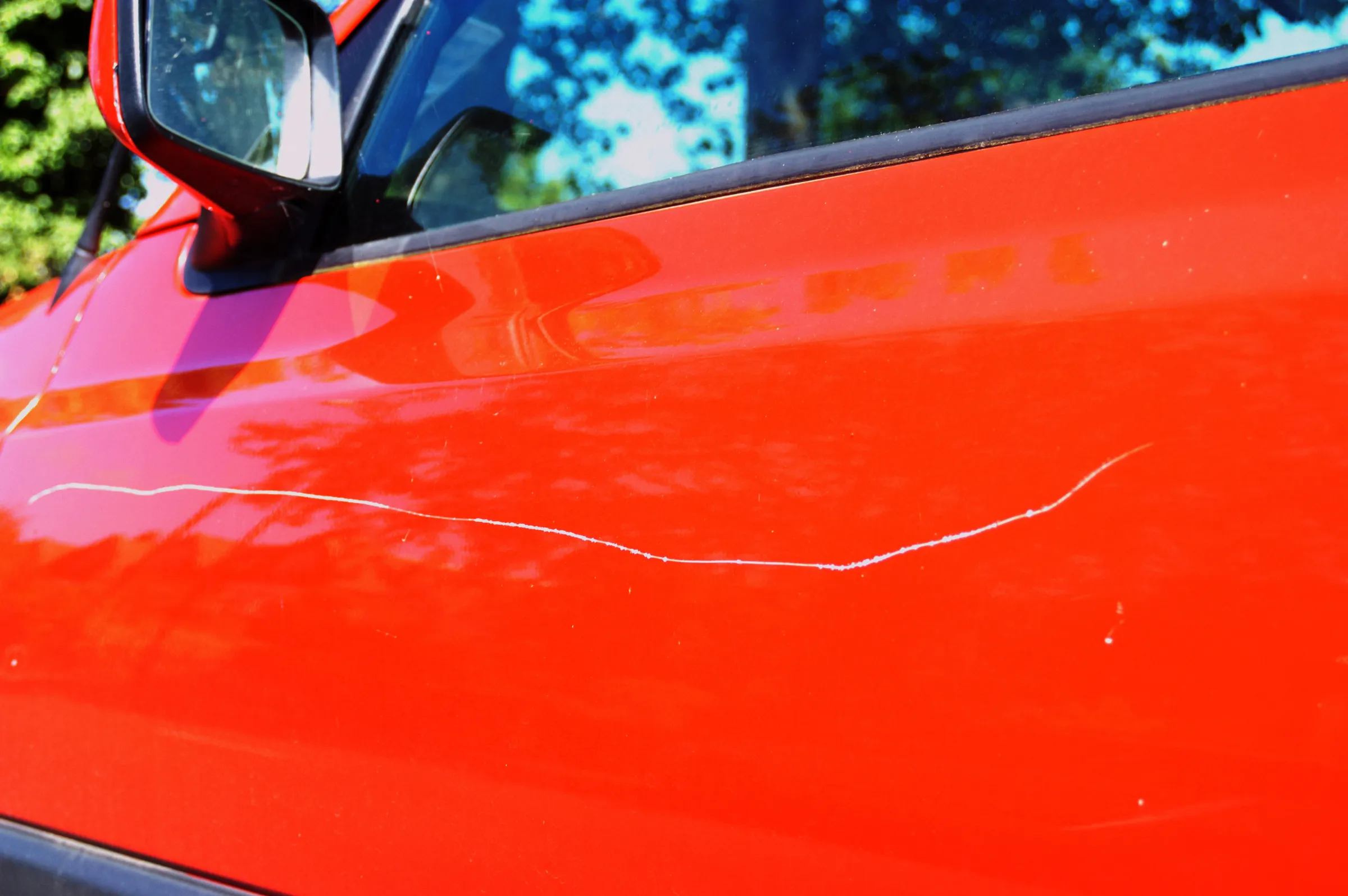 Close-up of a visible scratch on the door of a red car, highlighting the need for scratch removal.