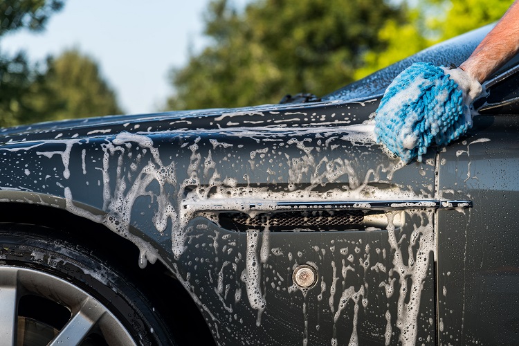 Hand washing a car with a blue microfiber mitt, covered in foam and soap suds.