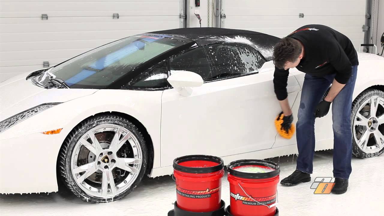 Person hand washing a white car with foam and two red buckets in the foreground.