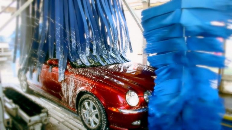 Red car being cleaned in an automatic car wash with blue brushes.