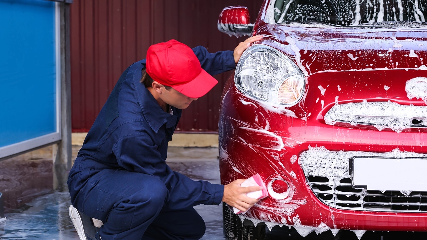 Man performing a hand car wash on a red car using a sponge and soap.