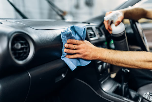 Professional car cleaner wiping the dashboard with a blue microfiber cloth and spray bottle.