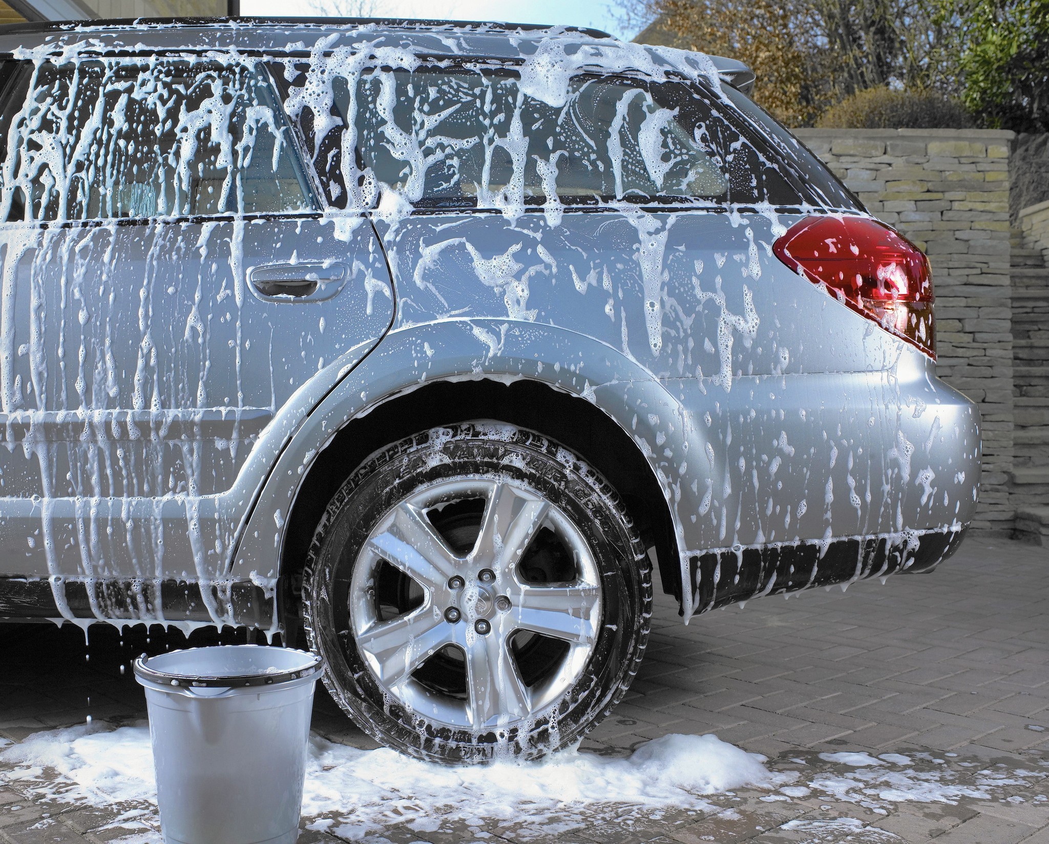 Home car wash with soap suds covering a silver car, bucket in foreground