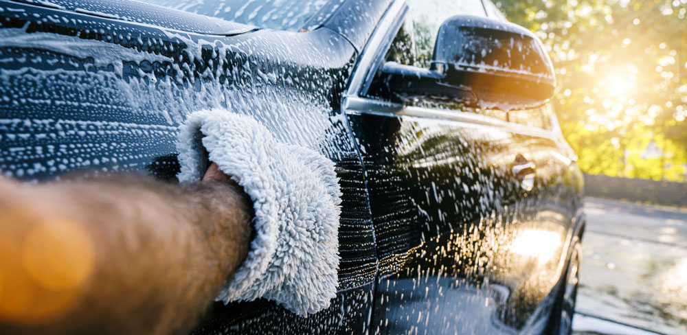 Close-up of a hand using a car wash mitt to clean a black car covered in suds.