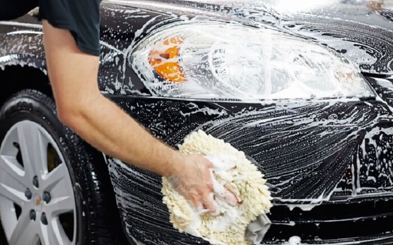 Person washing a car's front panel and headlight with a wash mitt during a fast hand car wash in the UK.