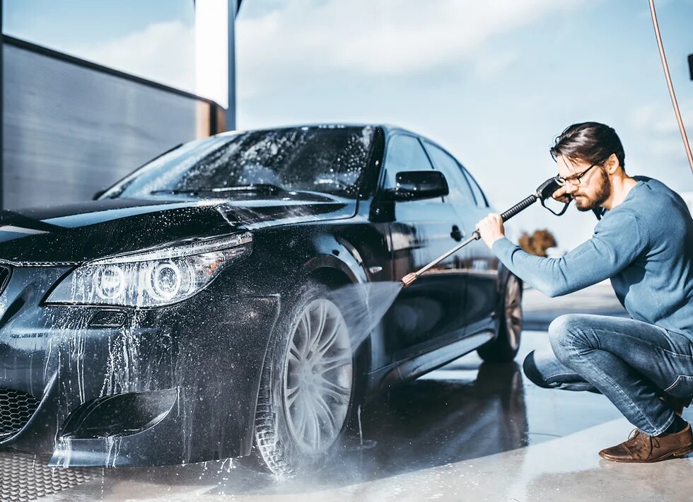 Man using a jet wash to clean a black car, focusing on the front area with water spray.