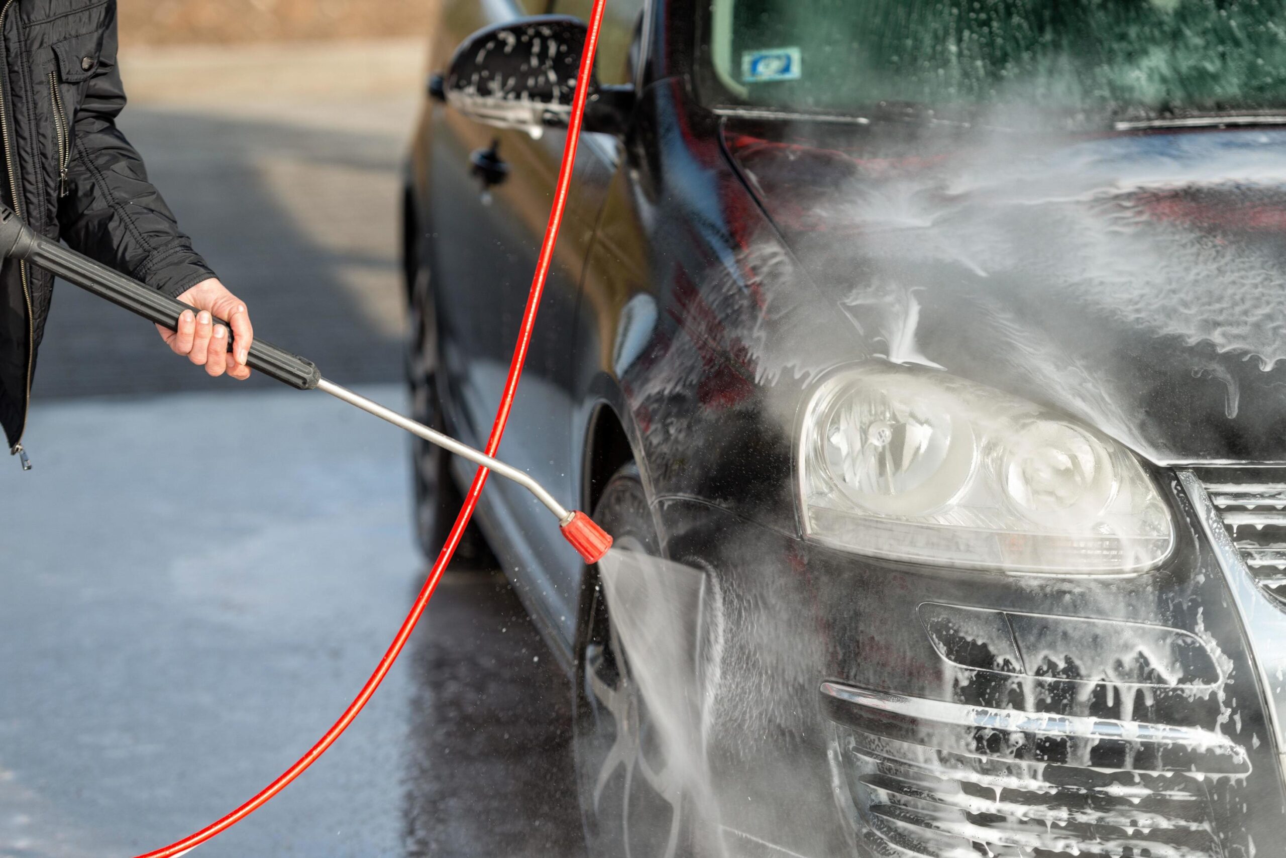 Person using a jet wash to clean a black car, focusing on the front headlight area.