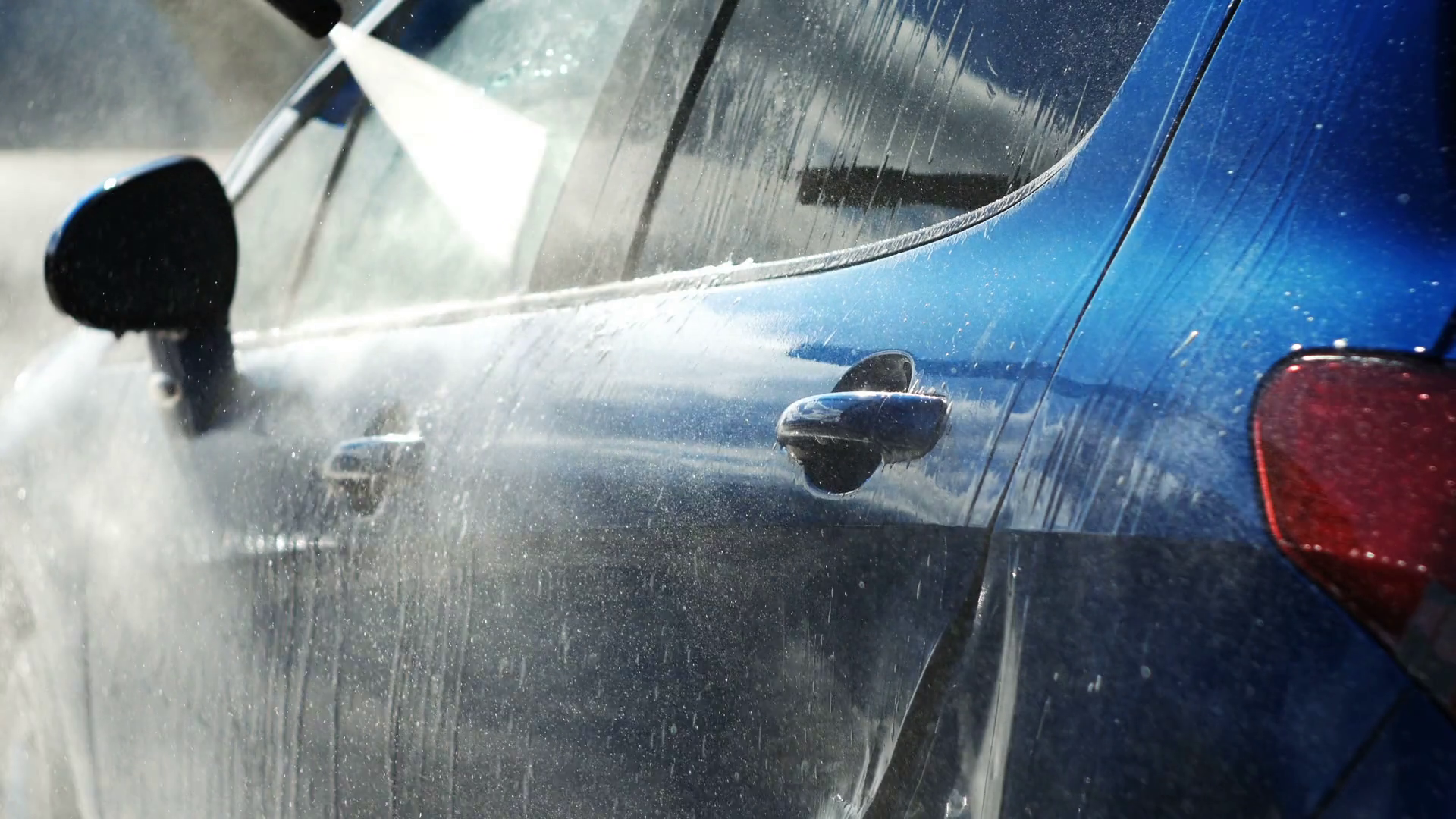 High-pressure water spray from a hose cleaning the side of a blue car.