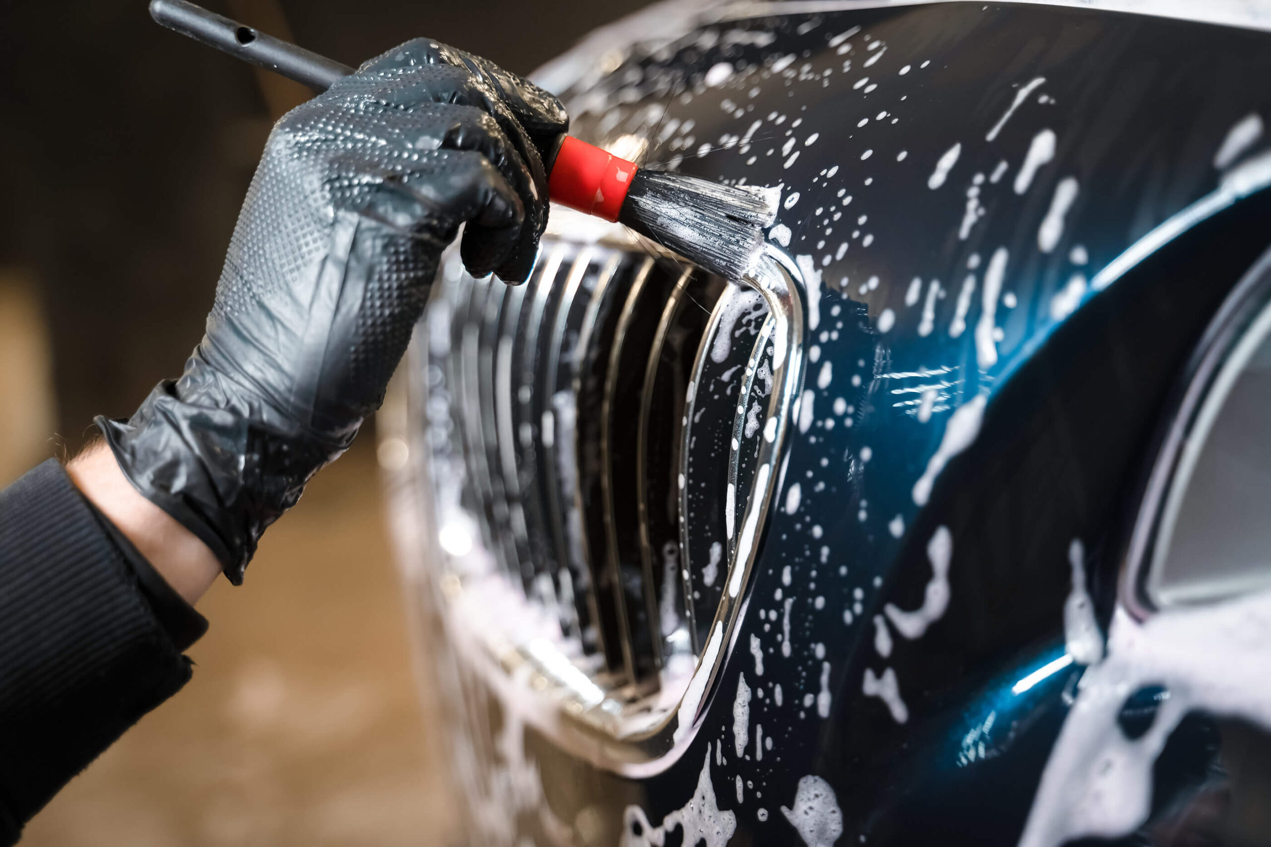 Close-up of a professional hand washing a luxury car's grille with a brush, ensuring meticulous care and protection.