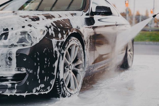 Black car being thoroughly washed with soap and water under high pressure, emphasizing proper car care.